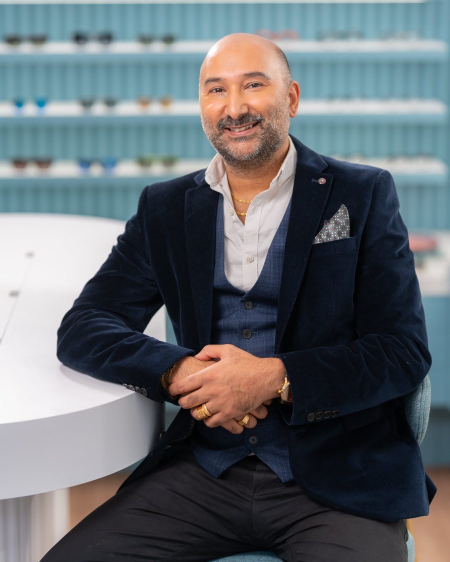 Mitesh Gandhi, smiling, seated in his Cambridge practice in a navy velvet jacket